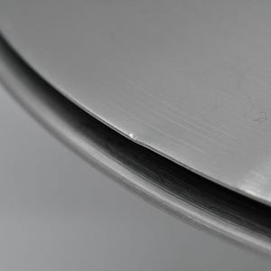 May include: Close-up of a metallic, circular object with a brushed finish. The edge of the object is visible, showing a slight curve and a darker, shadowed area. The surface has subtle imperfections.