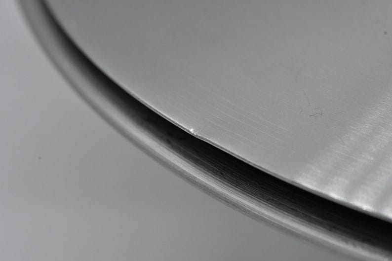 May include: Close-up of a metallic, circular object with a brushed finish. The edge of the object is visible, showing a slight curve and a darker, shadowed area. The surface has subtle imperfections.