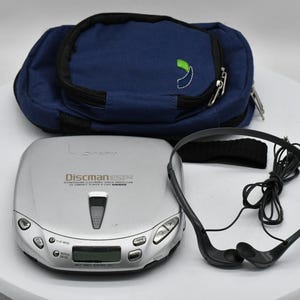 May include: A silver Discman ESP CD player with a blue carrying case and black headphones. The Discman has a digital display and control buttons. The carrying case has a green and white logo.