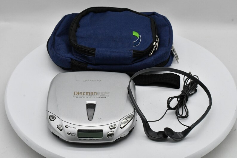 May include: A silver Discman ESP CD player with a blue carrying case and black headphones. The Discman has a digital display and control buttons. The carrying case has a green and white logo.