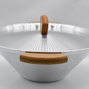 May include: A silver-colored wok with a wooden handle and two wooden side handles. The lid has a wooden handle and a ridged design. The wok is set against a white background.