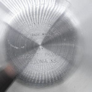 May include: Close-up of a shiny, silver metal surface with a circular pattern. The text "ATOM-POP TRADE MARK" is stamped in the center, with "BY JUNCRAFT PRODUCTS FREDONIA, KS" below. The surface has a brushed texture.