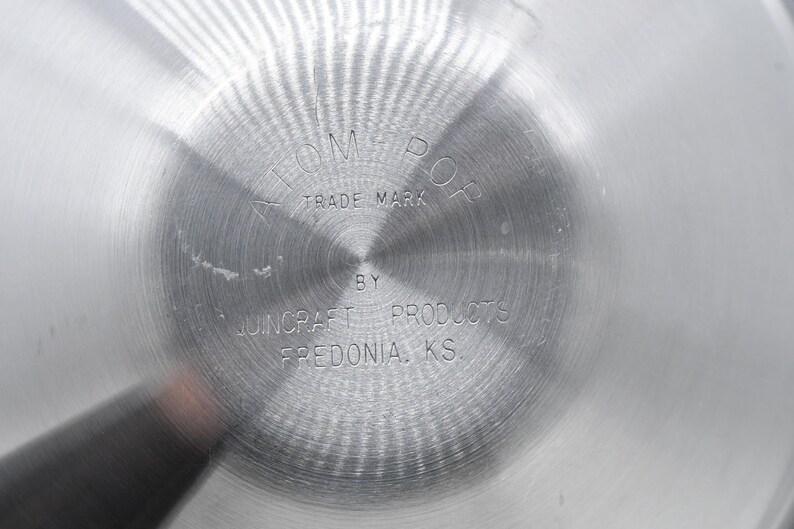 May include: Close-up of a shiny, silver metal surface with a circular pattern. The text "ATOM-POP TRADE MARK" is stamped in the center, with "BY JUNCRAFT PRODUCTS FREDONIA, KS" below. The surface has a brushed texture.