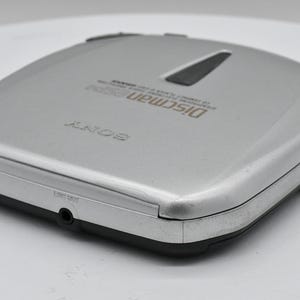 May include: Silver Sony Discman portable CD player. The device has a rectangular shape with rounded corners and a black trim. The top of the player has the word "SONY" and "Discman" printed on it. The player has a headphone jack and other control buttons.