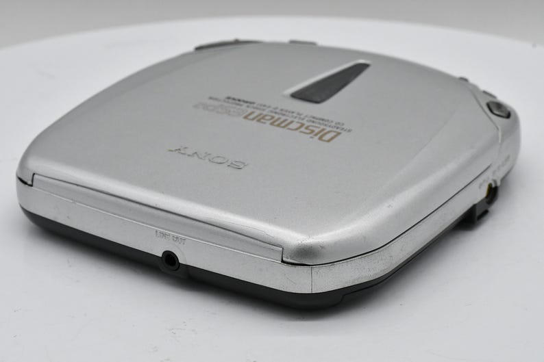 May include: Silver Sony Discman portable CD player. The device has a rectangular shape with rounded corners and a black trim. The top of the player has the word "SONY" and "Discman" printed on it. The player has a headphone jack and other control buttons.