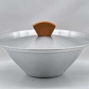 May include: A silver-colored wok with a wooden handle on each side and a wooden handle on the lid. The wok has a wide, rounded shape and is designed for cooking. The lid has a fan-shaped wooden handle.
