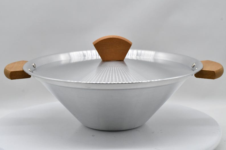 May include: A silver-colored wok with a wooden handle on each side and a wooden handle on the lid. The wok has a wide, rounded shape and is designed for cooking. The lid has a fan-shaped wooden handle.