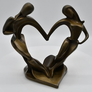 May include: A bronze-colored sculpture depicting two abstract human figures standing on a shared base. The figures are positioned to form a heart shape with their bodies, with their arms reaching upward and hands touching at the top center point.