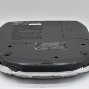 May include: A black and silver Sony portable CD player, viewed from the bottom. The device has a rounded rectangular shape with a silver trim and black buttons. The bottom surface has several labels and a battery compartment.