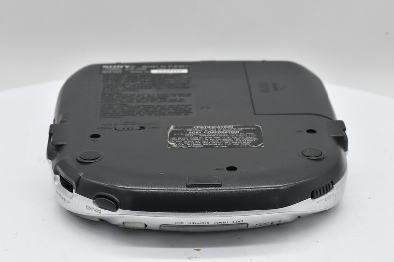 May include: A black and silver Sony portable CD player, viewed from the bottom. The device has a rounded rectangular shape with a silver trim and black buttons. The bottom surface has several labels and a battery compartment.