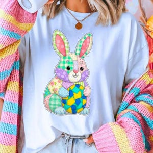 May include: White t-shirt featuring a colorful patchwork bunny holding a patterned egg. The bunny design includes pink, green, and purple hues. A multi-colored striped cardigan in pink, yellow, and blue is draped over the shoulders. The theme is Easter.