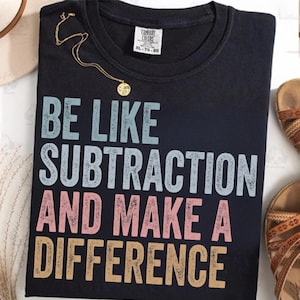 以下が含まれることがあります： 「BE LIKE SUBTRACTION AND MAKE A DIFFERENCE」の文字が青、ピンク、金色の黒いTシャツ。ペンダント付きのゴールドネックレスがシャツの上にかけられています。背景にはサンダルが見えます。