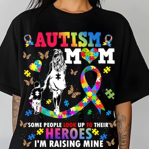 Op de afbeelding: Zwart T-shirt met een kleurrijk autisme-lint en de tekst "Autism Mom". Het ontwerp bevat ook een hart gemaakt van puzzelstukjes en de tekst "Sommige mensen kijken op naar hun helden, ik voed de mijne op."