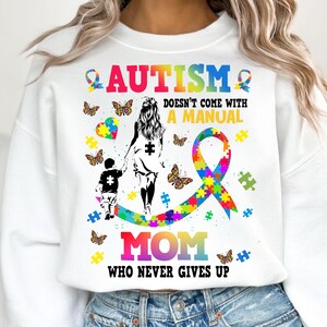 May include: White sweatshirt with a colorful autism awareness ribbon, puzzle pieces, and butterflies. The text reads "Autism doesn't come with a manual Mom who never gives up."