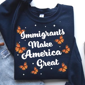 Puede incluir: Una sudadera azul marino con el texto "Immigrants Make America Great" en letras blancas. La sudadera está decorada con gráficos de mariposas naranjas.