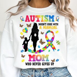 Op de afbeelding: Wit sweatshirt met een kleurrijk autisme-bewustzijnslint, puzzelstukjes en vlinders. De tekst luidt "Autisme komt niet met een handleiding. Moeder die nooit opgeeft."