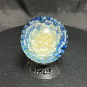 May include: A glass marble with a floral design in shades of blue and yellow. The marble sits on a black stand with the word "Glass" embossed on it. The marble's design resembles a flower.