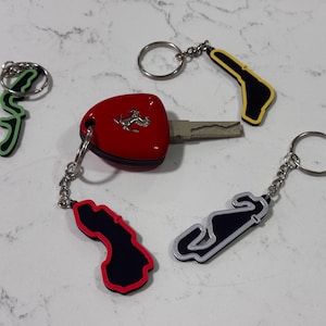 May include: Four keychains featuring different race tracks. One keychain has a red car key fob with a Ferrari logo. The other three keychains are black, white, and yellow with black outlines.