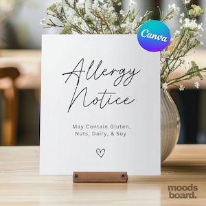 Pode incluir: Um letreiro branco com as palavras "Allergy Notice" em escrita elegante. Abaixo, diz "May Contain Gluten, Nuts, Dairy, & Soy". Um pequeno coração está na parte inferior. O letreiro é segurado por uma base de madeira, com flores em um vaso atrás.