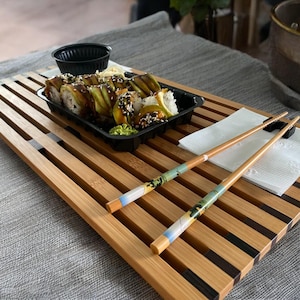 Puede incluir: Una bandeja de servir de madera con detalles negros, que presenta un plato de rollos de sushi y un par de palillos sobre una servilleta de papel blanca.