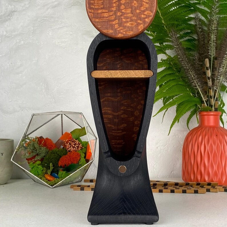 Op de afbeelding: Een hoge, verticale houten displaystandaard met een zwart, gebogen frame, een donker, generfd houten interieur en &eacute;&eacute;n horizontale plank. Bovenop bevindt zich een rond, roodbruin houten accentstuk en een kleine metalen stip is zichtbaar.