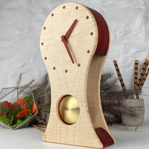 Può includere: Un orologio in legno con un quadrante colorato chiaro e un retro e una base rosso scuro. L'orologio ha un pendolo in ottone e lancette rosso scuro. Il quadrante ha piccoli punti marrone scuro che segnano le ore. È un oggetto decorativo.