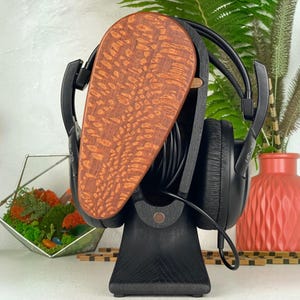 Op de afbeelding: Een zwarte houten hoofdtelefoonstandaard met een druppelvormig voorpaneel met een natuurlijk roodbruin houtnerfpatroon. De standaard houdt een zwarte over-ear hoofdtelefoon vast met een zwarte kabel opgerold in het open middenframe.