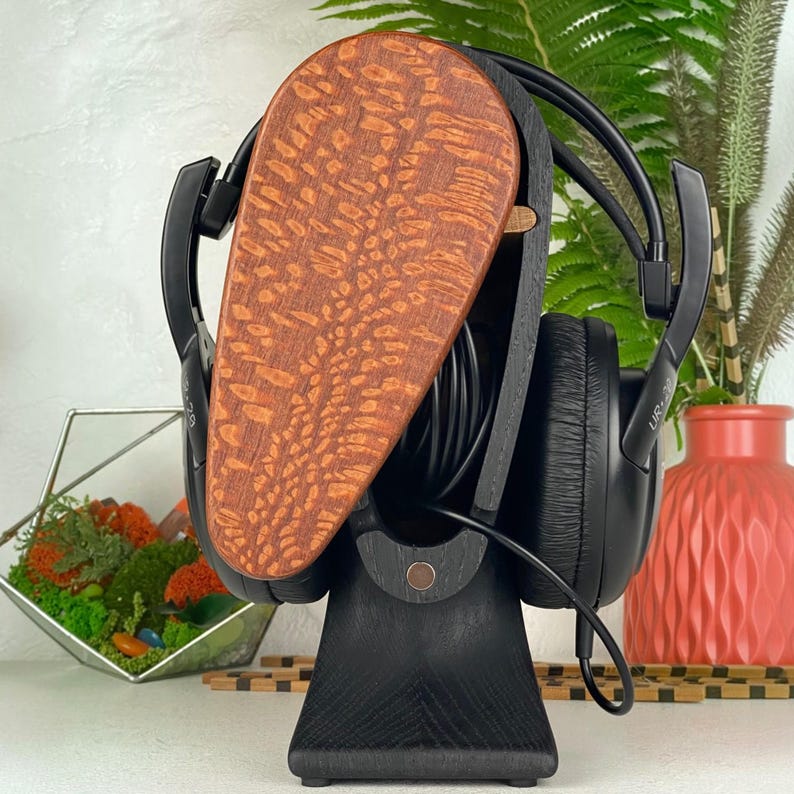Op de afbeelding: Een zwarte houten hoofdtelefoonstandaard met een druppelvormig voorpaneel met een natuurlijk roodbruin houtnerfpatroon. De standaard houdt een zwarte over-ear hoofdtelefoon vast met een zwarte kabel opgerold in het open middenframe.