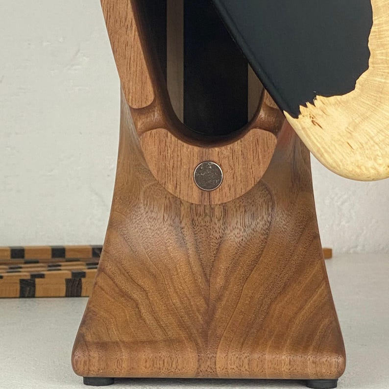 May include: A vertical wooden knife block featuring a smooth, light brown grain with a central circular metal accent. A portion of a black and light wood knife handle is visible, extending from the top opening of the block structure.