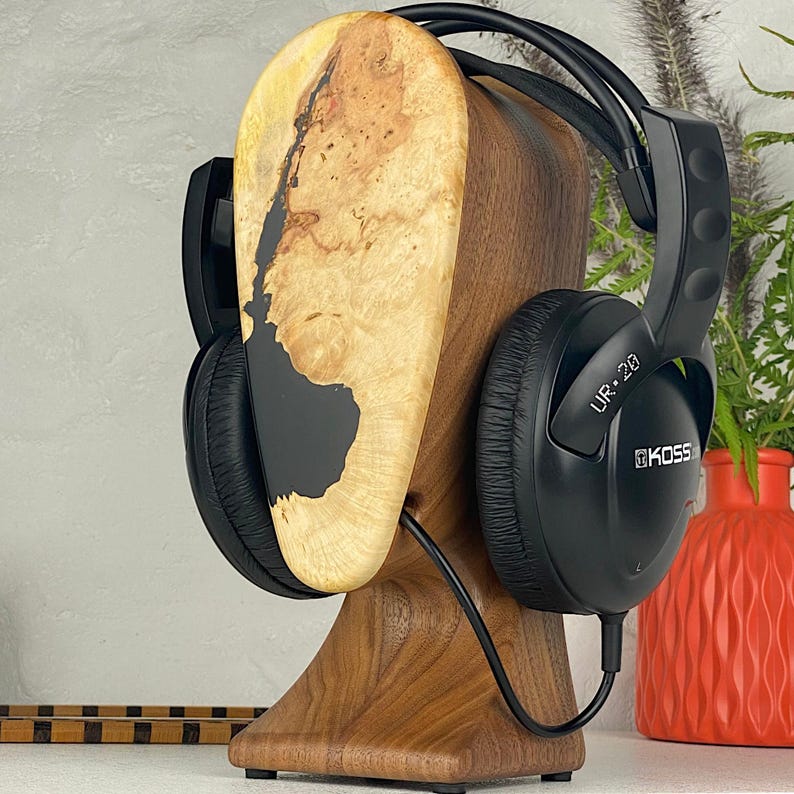 May include: A wooden headphone stand featuring a natural burl wood face with a black resin inlay, supporting a pair of black Koss UR-20 wired over-ear headphones. The stand has a smooth, dark walnut-toned base and a curved, upright display panel.