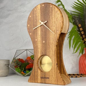 May include: A wooden mantel clock with a distinct grain pattern, featuring a circular face with light-colored hour markers, thin metallic hands, and a circular brass pendulum visible through a lower cutout. The brand name KAZMIDI is printed at the base.