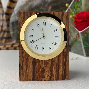 Puede incluir: Un reloj de escritorio de madera con un bisel dorado y una esfera blanca. La esfera del reloj presenta números romanos negros y la palabra "QUARTZ". La base de madera tiene un patrón marrón a rayas.