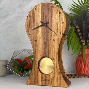 Può includere: Un orologio da tavolo in legno con venature verticali, caratterizzato da un quadrante rotondo con indicatori delle ore scuri e sottili lancette nere. Un pendolo circolare in ottone è visibile nella parte inferiore. La parola Hazmidi è stampata alla base.