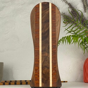 May include: A tall, vertically oriented wooden serving board with a rounded top and an hourglass-shaped silhouette. The board features a central dark brown walnut panel flanked by two light maple strips and outer panels of reddish-brown wood.