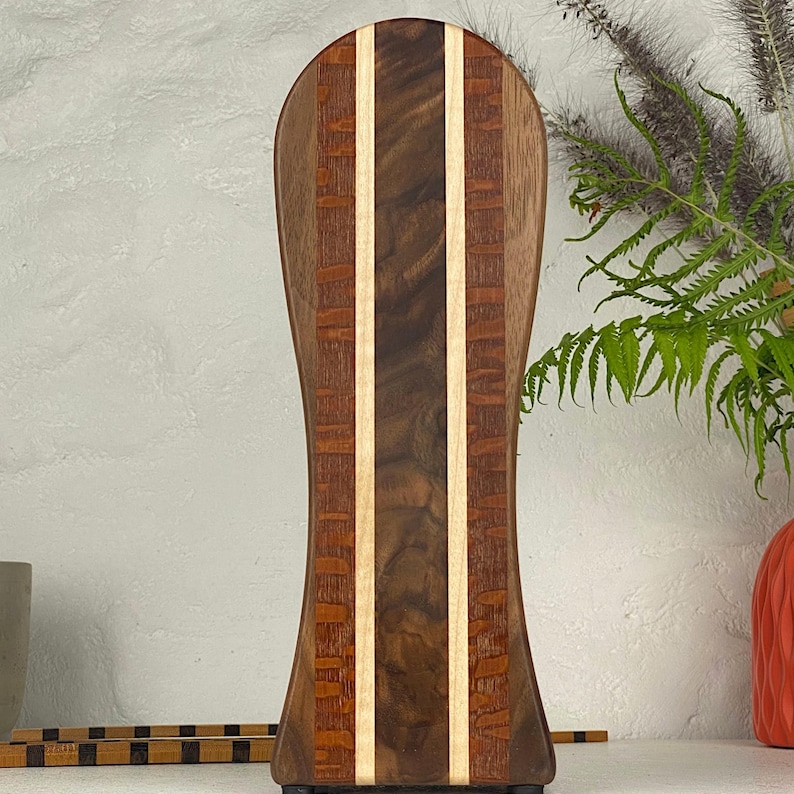 May include: A tall, vertically oriented wooden serving board with a rounded top and an hourglass-shaped silhouette. The board features a central dark brown walnut panel flanked by two light maple strips and outer panels of reddish-brown wood.