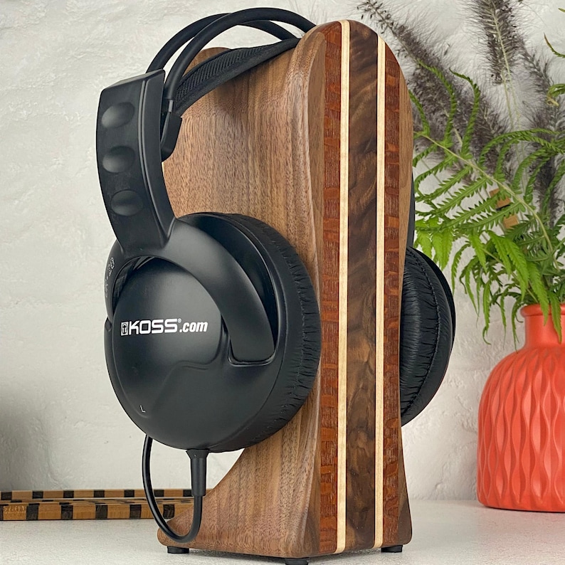 May include: Black over-ear headphones with the text KOSS.com printed on the side, resting on a vertical wooden stand. The stand features a dark brown wood grain with two vertical light-coloured wood inlay stripes running down the centre.