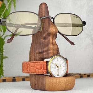 May include: A wooden eyeglasses and watch holder with a brown finish. The holder displays a pair of black-framed eyeglasses and a gold-toned watch with a white face and a brown leather band. The watch is a Tissot.