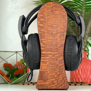 Op de afbeelding: Een zwarte over-ear hoofdtelefoon rustend op een verticale houten standaard met een opvallend golvend nerfpatroon. De hoofdtelefoon heeft gewatteerde oorschelpen en een flexibele hoofdband, centraal geplaatst op de gladde houten houder.