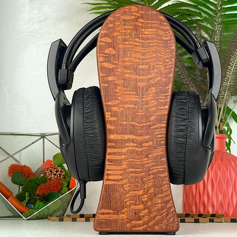 Op de afbeelding: Een zwarte over-ear hoofdtelefoon rustend op een verticale houten standaard met een opvallend golvend nerfpatroon. De hoofdtelefoon heeft gewatteerde oorschelpen en een flexibele hoofdband, centraal geplaatst op de gladde houten houder.
