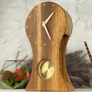 Puede incluir: Un reloj de madera con un diseño único. El reloj presenta un patrón de rayas marrón claro y marrón oscuro, con un elemento circular de color latón. Las manecillas del reloj son de color claro y el reloj tiene una forma curva.