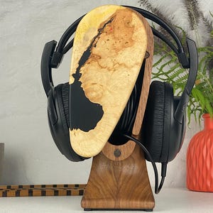 May include: A pair of black over-ear headphones resting on a wooden stand. The stand features a vertical support base made of dark brown wood and a top section composed of light-coloured burl wood with a smooth, black resin inlay.