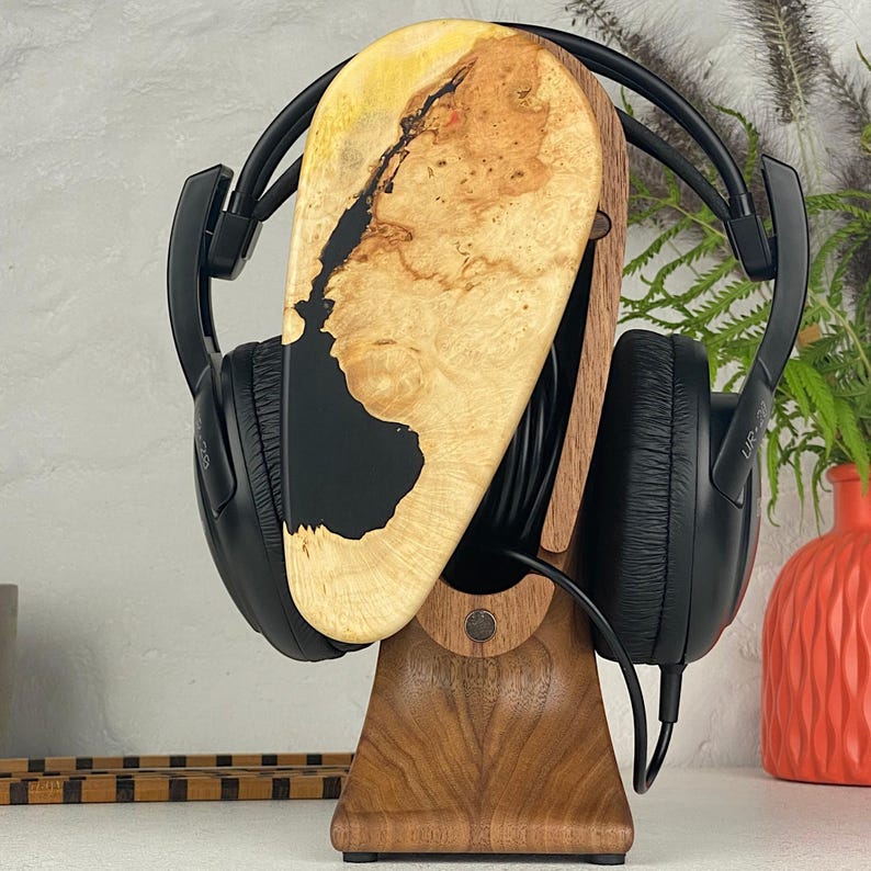 May include: A pair of black over-ear headphones resting on a wooden stand. The stand features a vertical support base made of dark brown wood and a top section composed of light-coloured burl wood with a smooth, black resin inlay.