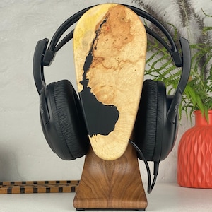 May include: A pair of black over-ear headphones resting on a wooden stand. The stand features a vertical, teardrop-shaped piece of light-coloured burl wood with a dark black resin inlay, supported by a solid, dark-toned wooden base.