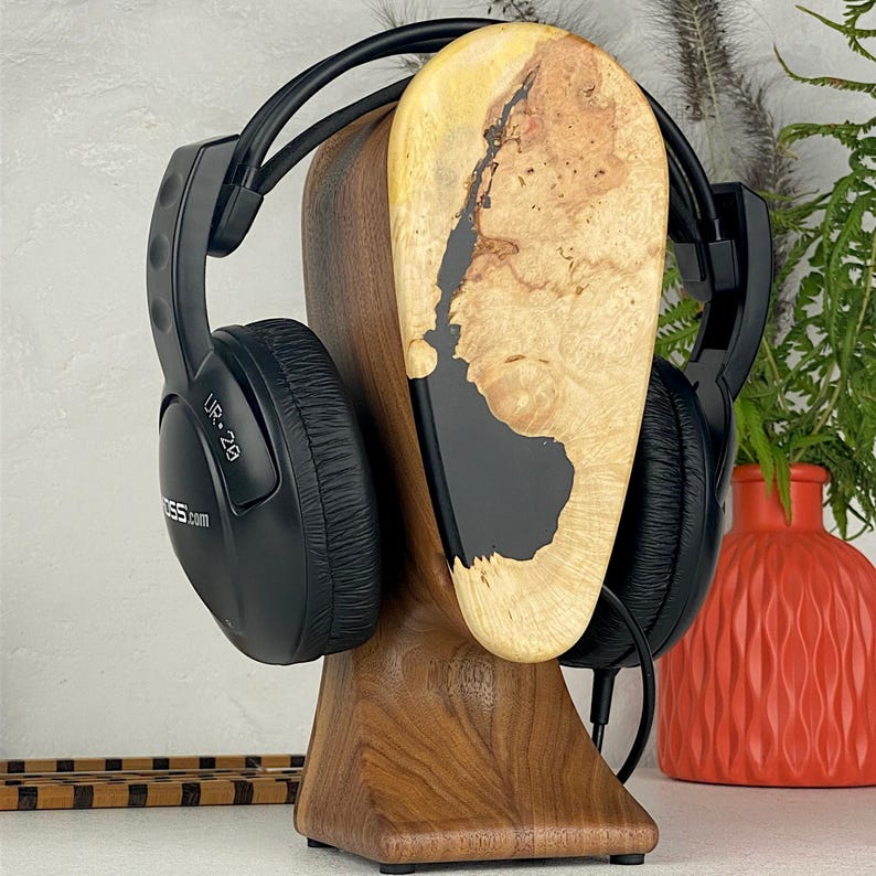 May include: A pair of black Koss UR-20 over-ear headphones resting on a wooden headphone stand. The stand features a dark walnut base and a light-coloured burl wood top section with a prominent black resin inlay running through the centre.