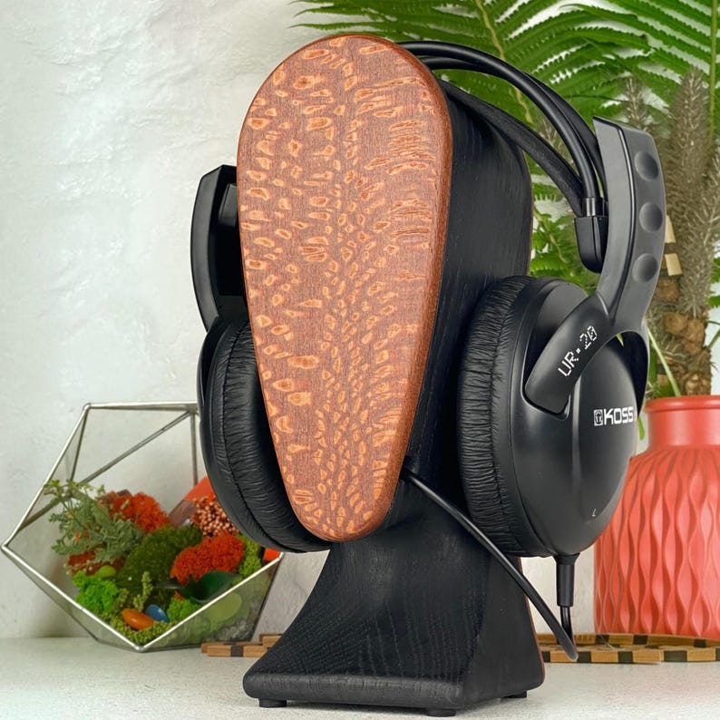 Op de afbeelding: Zwarte Koss UR-20 hoofdtelefoon rustend op een gestileerde houten standaard. De standaard heeft een donkerzwarte voet en een verticaal, druppelvormig voorpaneel met een natuurlijk, lichtbruin houtnerfpatroon. De hoofdtelefoon heeft zwarte oorschelpen.