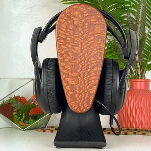 Op de afbeelding: Zwarte over-ear hoofdtelefoon op een standaard met een gladde, zwarte voet en een verticaal, ovaal houten paneel met een opvallende, textuurrijke, lichtbruine houtnerf. De hoofdtelefoon is voorzien van zwarte oorkussens.