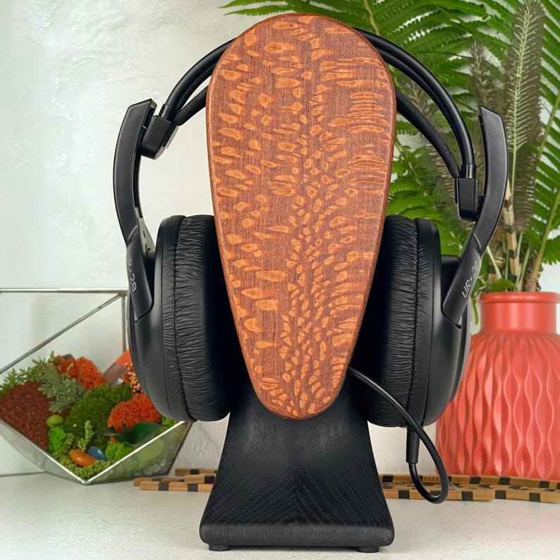 Op de afbeelding: Zwarte over-ear hoofdtelefoon op een standaard met een gladde, zwarte voet en een verticaal, ovaal houten paneel met een opvallende, textuurrijke, lichtbruine houtnerf. De hoofdtelefoon is voorzien van zwarte oorkussens.