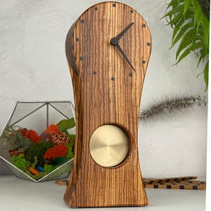 May include: A tall, arched wooden mantel clock featuring a prominent vertical wood grain pattern, dark brown hour and minute hands, and small dark circular hour markers. A circular, brushed brass pendulum disc is visible in the lower section.