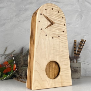 Op de afbeelding: Een houten bureauklok met een lichtgekleurde, gewelfde voorkant en een donkere houten basis. De klok heeft een ronde opening en houten wijzers. De wijzerplaat van de klok heeft kleine, donkere stippen die de uren markeren. De klok is ongeveer 25 cm hoog.