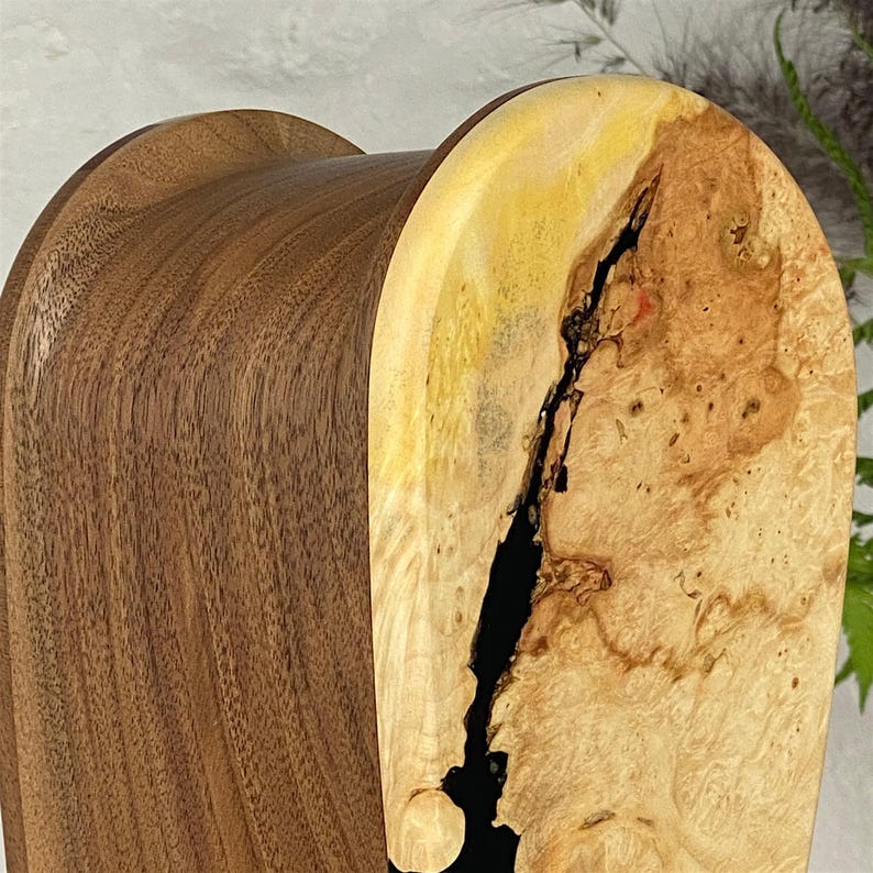 May include: A curved wooden object featuring a smooth, dark brown walnut grain on the side and a light-coloured, textured burl wood face. A deep, vertical black resin inlay runs through the centre of the burl wood, creating a striking contrast.